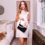 Victoria Magrath stands next to a window an a large metal radiator in a bedroom at home. She wears a sleeveless silver dress and holds a black leather and silver stud handbag.