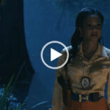 Christina Milian is seen looking to one side at night time. She stands in front of a lake on a grassy verge. Christina wears a shiny long sleeve blouse, large belt and dark orange trousers.