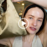Victoria Magrath holds up a dark gold bag. She wears a black hair band to push back her loose hair. Victoria appears in her attic filming room at home with no makeup on.