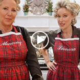 Leonora Smee stands next to her mother in the kitchen of her countryside home. Leonora wears her hair up in rollers and is seen in a dark green silk shirt dress, a tartan style apron and pearl jewellery.