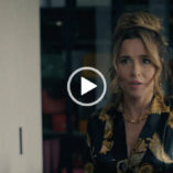 Linda Cardellini is seen wearing her hair up in a large bun. She appears in a black and gold print wrap nightwear top. Linda stands in a dressing room in front of large windows in partial darkness.