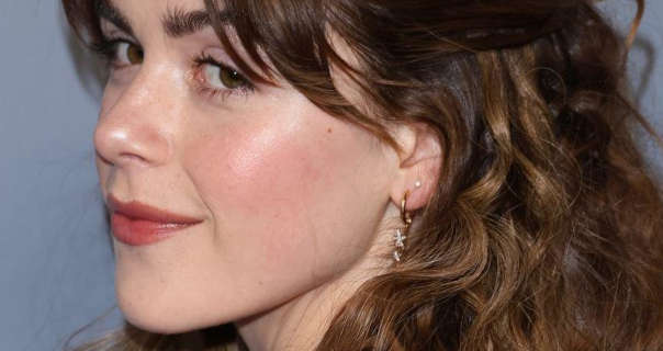 Kiernan Shipka is seen front the side as she turns and looks at the camera. She stands in front of a grey gradient wall. Kiernan has her brown hair down and in curls. She appears in a dark red lipstick, black mascara and a pair of hanging gold hoop and star earrings.