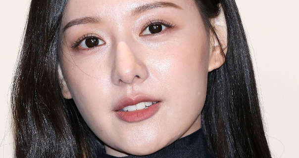 Kim Ji-won tilts her head slightly. She stands in front of a dark cream wall with her black hair down and straightened. Kim Ji-won wears a dark pink lipstick with black lash defining mascara. She is seen in a high neck black dress.