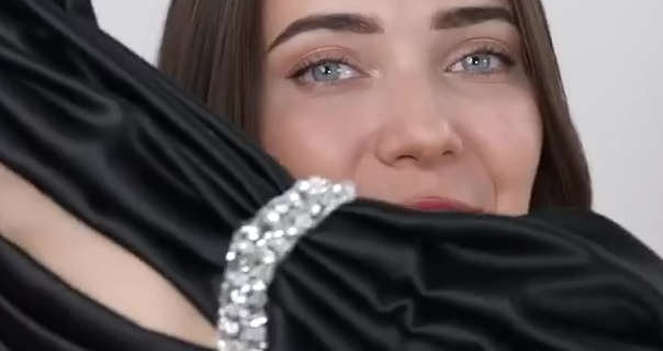 Roxxsaurus sits in front of an off-white wall. She has her long brown hair straightened and down. Roxi holds up a black and gemstone evening gown during one of her try on fashion hauls for YouTube.