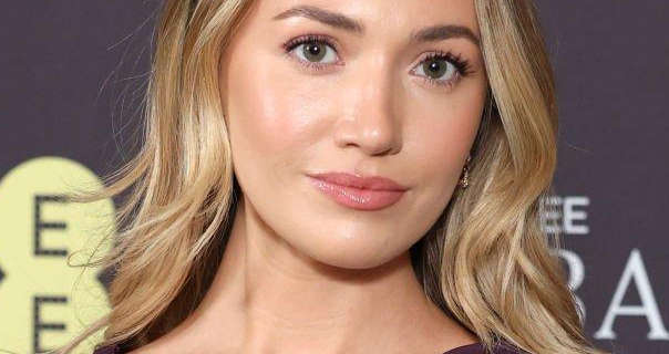 Tilly Keeper appears in front of a dark grey wall with a yellow logo and white text on it. She has her long blonde hair down and in natural waves. Tilly wears a pink gloss lipstick, black mascara and a pair of dainty gold earrings.