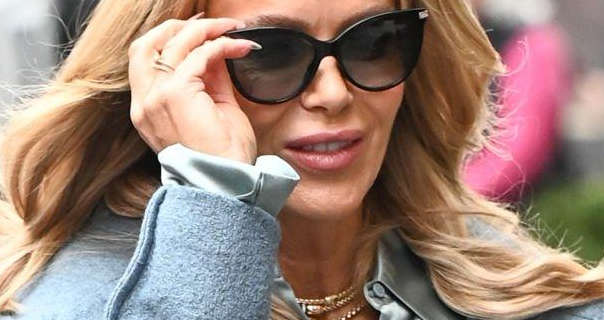 Amanda Holden touches the thin black frame of the sunglasses she is wearing with one hand. She walks with her long blonde hair loose and blowing a little in the wind. Amanda wears a bobbly powder blue coat over a shiny pastel blue blouse.