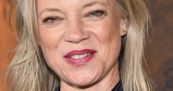 Amy Smart wears her dark blonde hair loose and a little messy. She stands in front of an orange and black backdrop. Amy is seen in a dark pink lipstick and soft black mascara.