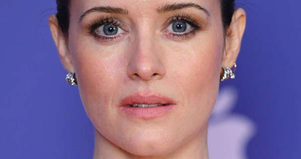 Claire Foy wears her hair tied up in a bun. She appears in front of a purple wall that has an out of focus white Apple logo on it. Claire has on a light pink lipstick, black mascara and a row of small silver circle earrings.