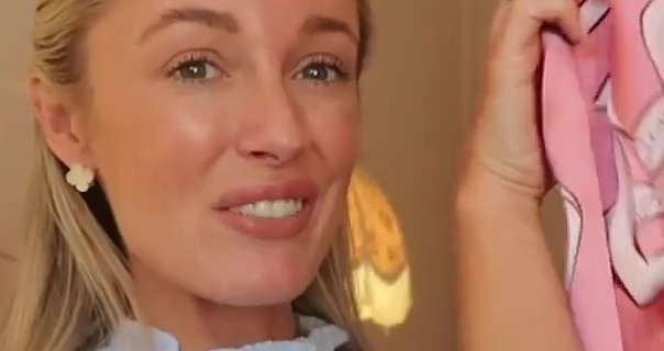 Josie Irons appears in one of her light brown bedrooms in front of a large yellow and orange lamp. She has her blonde hair down and pushed back. Josie holds up a pink silk Olivia Von Halle pyjama top. She is seen in a sheen lipstick, black mascara and a pair of small four petal design earrings.