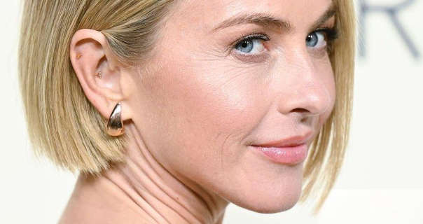 Julianne Hough appears in front of an off-white wall with thin black writing on it. She has her blonde hair cut short in a bob. Julianne wears a light pink gloss lipstick and a pair of chunky gold earrings. She is turning her head to look into the camera and is smiling a little.