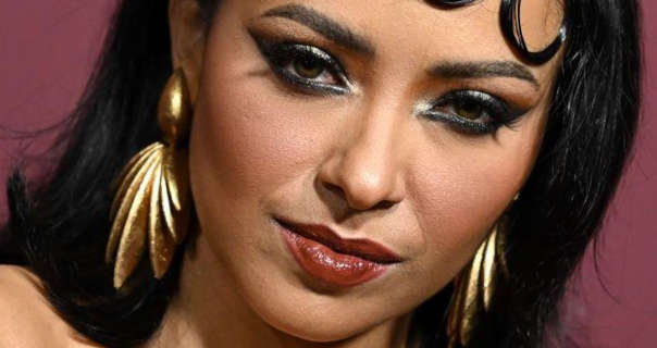 Kat Graham appears in front of a dark pastel red wall. She wears a pair of large gold sheet earrings with black winged eyeliner and a deep red lipstick. Kat wears her long black hair pushed back.