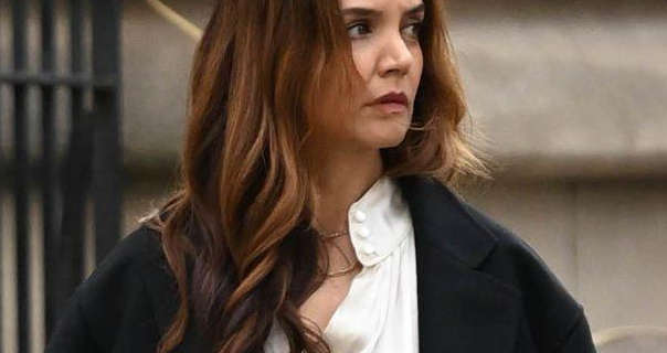 Katie Holmes looks to her left. She wears her thick brown hair loose and in soft waves. Katie is seen in a satin button blouse and a long black coat. She walks in front of a large stone building and black metal railings.