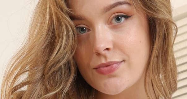 Kitty Cashew wears her brown and blonde hair loose and in waves. She appears in front of a cream wall and closed cream shutter blinds. Kitty stars in a dark pink lipstick and black winged eyeliner.