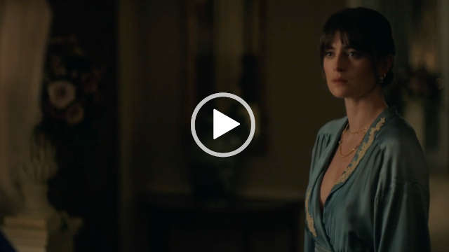 Millie Brady wears a dusty blue wrap over silk and ivory lace gown. She appears in a dark open room. Millie stars with her black hair tied back. She is seen in gold jewellery, a dark red lipstick and black mascara.