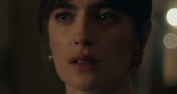 Millie Brady is seen in a dark room with a few light flares behind her. She wears her black hair tied back. Millie stars in a pair of dark gold earrings, black mascara and a dark red lipstick.