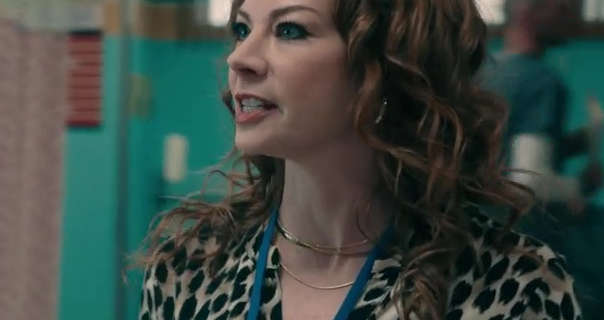 Katie Page appears in a hospital settings. She walks through a green, red and orange treatment area. Katie has her light brown hair in curls and is wearing an animal print top with a thin gold necklace and gold earrings.