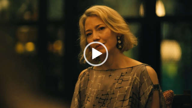 Carrie Coon sits in a restaurant in front of a row of full length black frame windows. She wears a dark olive cold shoulder satin top. Carrie is seen with her blonde hair cut short. She appears in a pair of long dangling earrings and is lit at nighttime by multiple lamps.