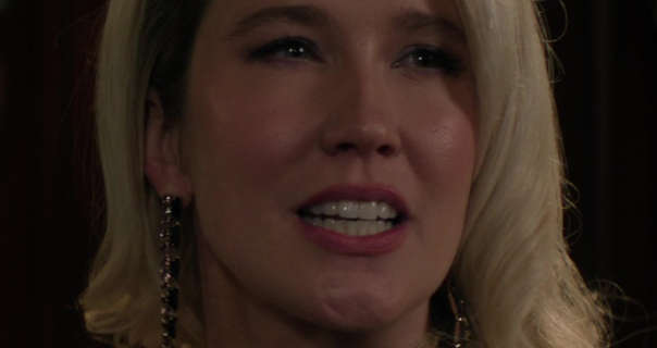 Anna Camp stands with her long blonde hair loose and curled. She wears a dark pink lipstick, black mascara and a pair of dangling gold jewel studded earrings. Anna is seen against a black backdrop looking slightly aggressive as she talks.