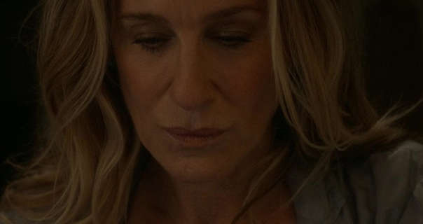 Sarah Jessica Parker is seen in the dark. She wears her long blonde hair down, loose and naturally messy. She is seen in. a dark pink lipstick, black mascara and a beige cardigan.