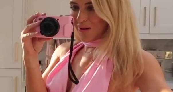 Em Sheldon records herself as she stands in her white cupboard and white marble London kitchen. She holds her pink vlog camera in one hand. Em wears a light pink halter neck gown with her blonde hair straightened and loose around her shoulders.