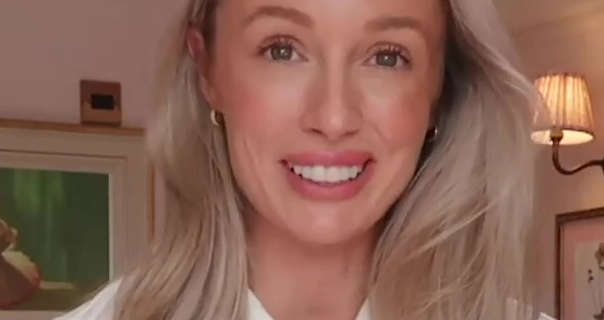 Josie Irons is seen standing in one of her living rooms at her home. She has her dark blonde hair loose and in soft waves. Josie wears a dark pink lipstick with a cream juimper and small gold circle earrings. She appears in front of two framed paintings and a small wall lamp.