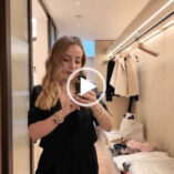 Fleur De Force stands in the wardrobe area of a room at The Emory in London. Fleur has her blonde hair loose and styled in waves. She wears a black cropped jacket with gold jewellery, a black camisole top and black trousers. Fleur records in the yellow light of the cream and light wood hotel room.