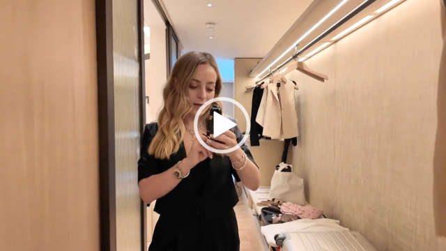Fleur De Force stands in the wardrobe area of a room at The Emory in London. Fleur has her blonde hair loose and styled in waves. She wears a black cropped jacket with gold jewellery, a black camisole top and black trousers. Fleur records in the yellow light of the cream and light wood hotel room.