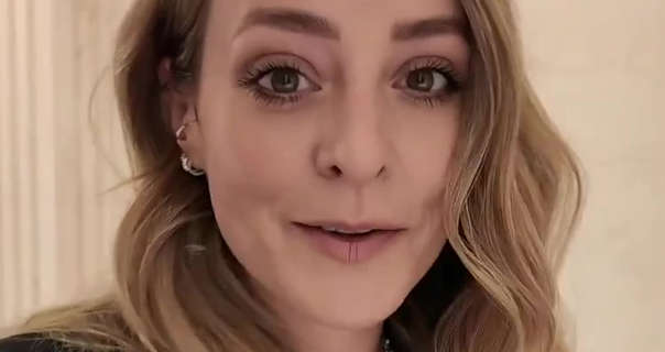 Fleur De Force is seen in front of a light neutral wall in a The Emory London hotel. Fleur has her long blonde hair in loose waves down around her shoulders. She is seen in Raven James earrings. She pairs a dark pink lipstick with black mascara and a matt foundation.