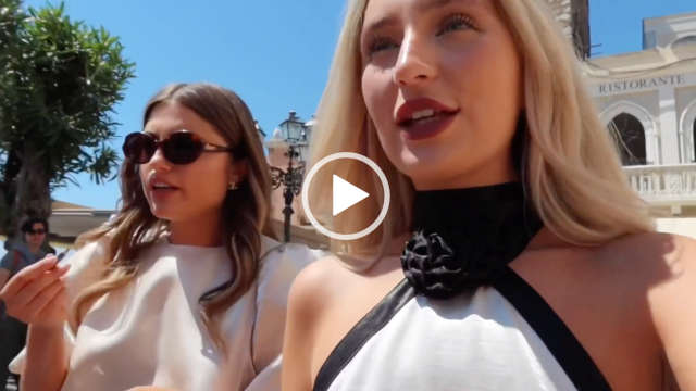Grace Shadrack and Grace Foley walk past classical buildings in Sicily on their way to a fashion shoot. Grace Foley wears a puffy champagne gold satin top with her long brown hair loose and in waves. She is seen in a pair of large sunglasses. Grace Foley has a lipstick pen in one hand as she touches up her makeup.