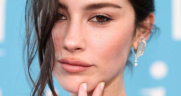 Gracie Abrams is seen in front of a light blue wall that has white text on it. She wears her black hair tied up with strands pulled down in front of one eye. Gracie is seen in a dark pink matt lipstick, black eyeliner, black mascara and a pair of large dangling silver earrings.