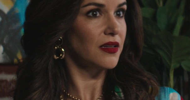 Melissa Fumero looks a little surprised as she sits at a table in front of a green plant and a blue, yellow and white abstract painting. Melissa has her black hair in waves. She stars in a dark red matt lipstick, black mascara, large gold earrings and a gold chain necklace.