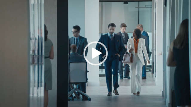 Aja Naomi King walks through a large open plan office. She wears a white blazer and trousers with an orange blouse. Aja has her black hair cut short. She carries a large light brown leather handbag.