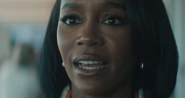 Aja Naomi King is seen with her black hair cut short. She wears a dark plum gloss lipstick with black mascara. Aja is seen in a cream blazer and orange top as she stands in an open plan office.