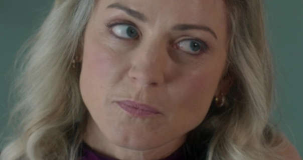 Aoife Mulholland stands in front of a grey green kitchen wall. She has her hair down and loose in soft waves. Aoife looks to one side. Her makeup is a dark pink matt lipstick, black eyeliner and curled eyelashes. She appears in a purple top and a pair of gold earrings.