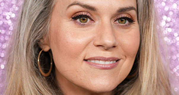 Hilarie Burton is seen in front of a light purple glitter poster wall. She has her long blonde with grey hair loose and down. Hilarie stars in a dark pink gloss lipstick, a pair of gold hooper earrings, dewy foundation and black mascara.