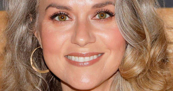 Hilarie Burton smiles as she is photographed in front of wood slat wall. She wears her grey blonde hair loose and curled. Hilarie appears in a gloss pink lipstick, black mascara and a pair of large thin gold hoop earrings.