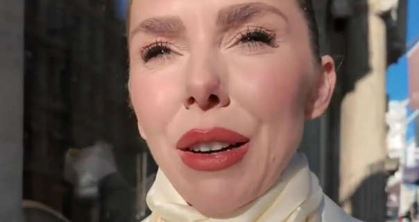 Laura Blair faces into the bright winter sunshine while standing in front of a shopfront street in London. She wears her hair tied up in a bun. Laura is seen in a cream silk scarf and a white blazer. She has a shiny red lipstick, matt foundation, heavy mascara and red blush for her makeup.
