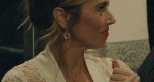 Linda Cardellini is seen from one side. She wears her brown hair tied up and with blonde highlights. Linda stars in a white silk trim and lace robe. She wears a dark pink lipstick with mascara and a pair of kite shape gemstone earrings.