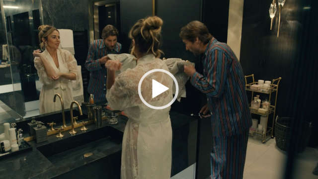 Linda Cardellini stands in front of a large mirror in a white and black stone bathroom. She applies a skin care lotion. Linda is seen with her hair tied up in a bun. She wears a long sheer white dressing gown.