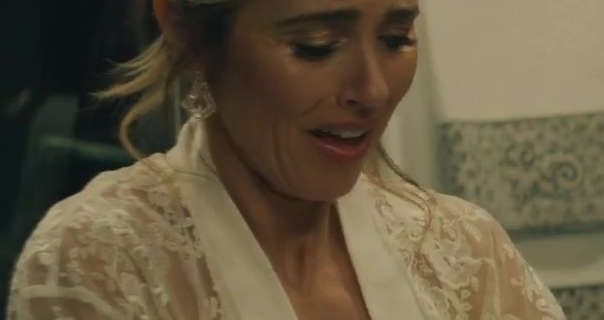 Linda Cardellini looks down while standing in a bathroom with cream towels hanging behind her. She has her hair tied up in a bun. Linda wears a sheer white robe with large gemstone earrings.