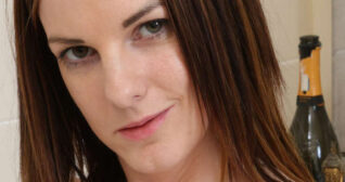 Isabella Rose smirks and looks into the camera. Her light brown shoulder length hair is down and straight. Isabella is seen in front of a cream bathroom tile wall. There is an open champagne bottle and small statue of Buddha behind her.