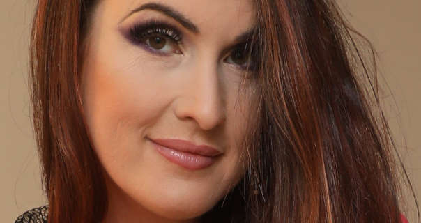 Petra V models in front of taupe wall. She has red highlights in her straight and loose brown hair. Petra is seen in a glossy dark pink lipstick and black mascara. She smiles slightly while looking directly into the camera.