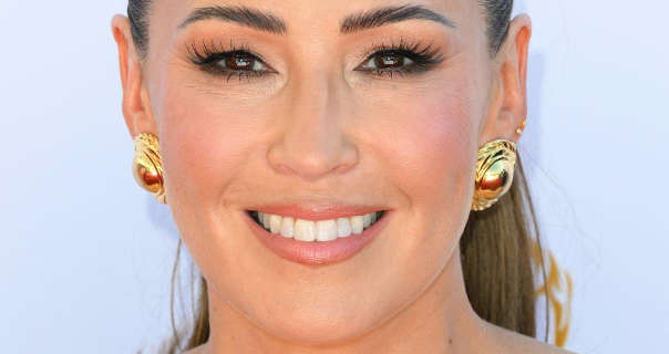 Rachel Stevens smiles while standing in front of an off-white wall. She wears a pair of large gold shell earrings with her light brown hair tied back in a ponytail. Rachel is seen in a dark pink lipstick, black eyeliner and black mascara.