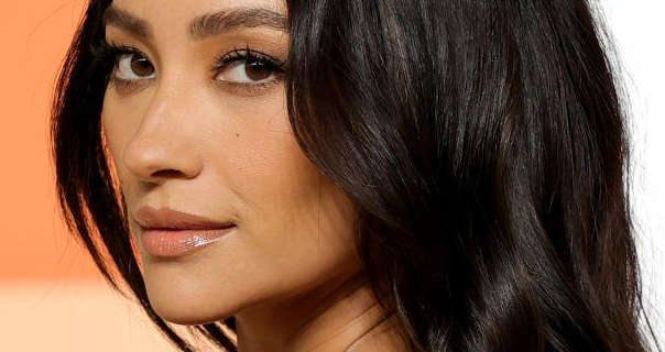 Shay Mitchell stands in front of a dark and pale orange wall. She has her long, thick black hair loose and in slight waves. Shay is seen in a dark pink gloss lipstick, black eyeliner and black mascara. She is pictured from the side, turning to look straight into the camera.