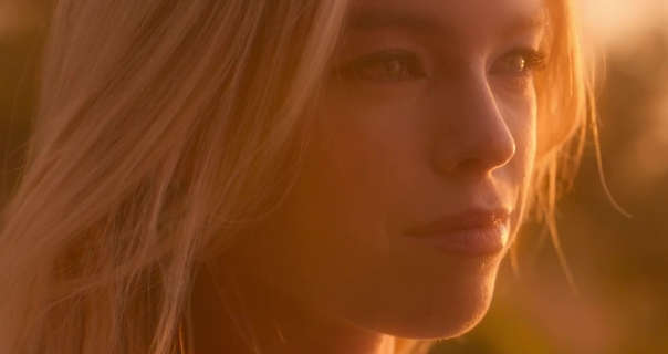 Milly Alcock is seen in the orange glow of the setting sun. She has her long blonde hair loose and blowing a little in the wind. Milly stars in a dark pink matt lipstick and black eyeliner.