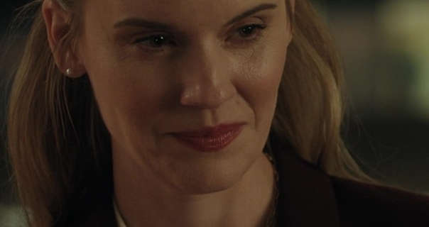 Maggie Grace appears in an office at night. She wears her blonde hair loose and pushed back. Maggie is seen in a dark red lipstick and peal earrings. She wears a dark maroon jacket over a cream top.
