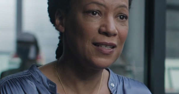 Nina Sosanya is seen in front of a black frame glass room in a large office. She wears her hair pinned back in tight braids. Nina stars in a dusty blue button up satin top, mauve lipstick with natural lashes. She wears a thin gold chain necklace in an episode of Surface.