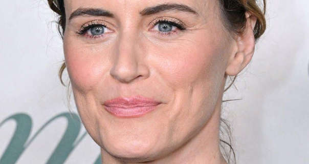 Taylor Schilling smiles and looks to the side. She stands in front of an off-white wall with dark green writing on it. Taylor is seen with her dark blonde hair tied up. She appears in a light pink matte lipstick, blush and black mascara.