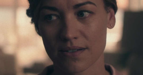 Yvonne Strahovski looks to one side. She sits in front of a set of large kitchen windows with the rising sun filtering through. Yvonne has her dark blonde hair pinned up in a bun. She wears a dark pink pyjama top, natural lashes, gem stud earrings and a dark pink lipstick.