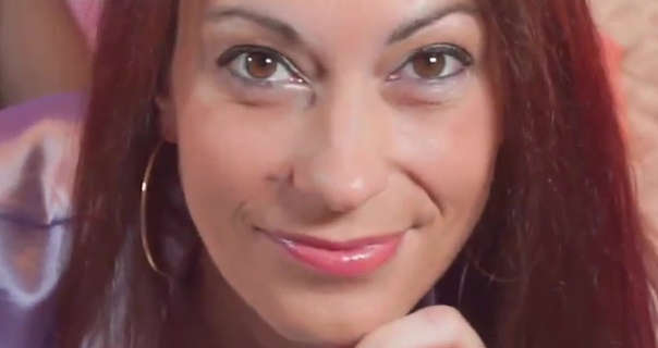 Vana looks into the camera during a Vanaweb OnlyFans livestream. She wears her thick brown hair loose and straight down. Vana lies on a bed covered in a gold quilted throw. She wears a shiny lilac top and black sheer stockings with a pink gloss lipstick, blue eyeliner and a pair of large thin gold hoop earrings.